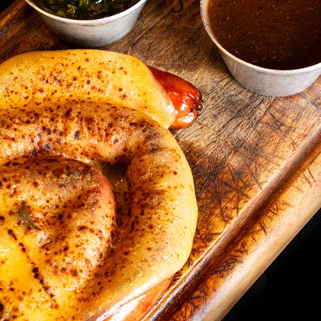 Linguiça defumada finalizada com queijo cremoso derretido, acompanhado de molho barbecue e salsa crioula.