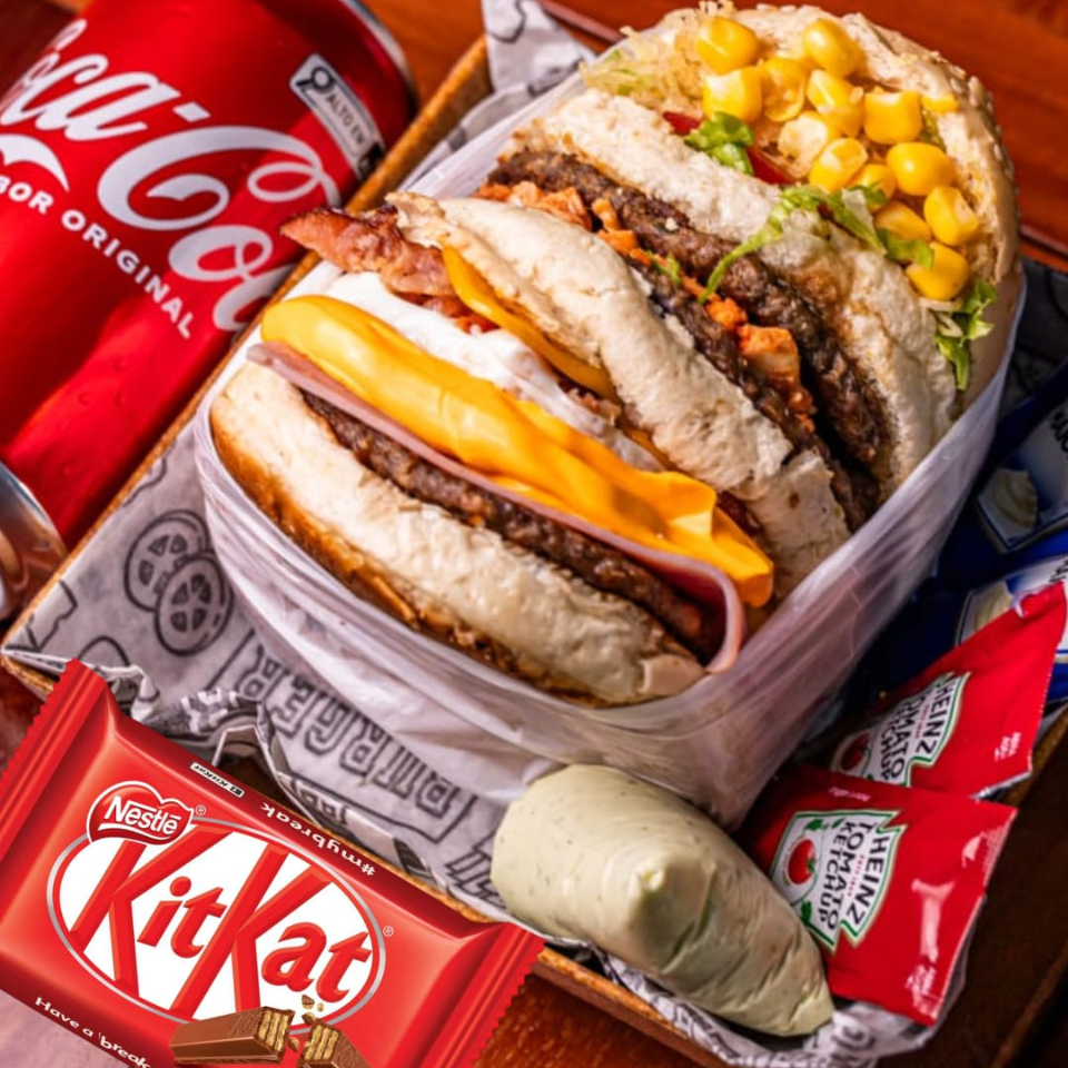 TRIPLO BURGUER ( Pão artesanal, 3 suculentas carnes de hambúrguer bovino, queijo prato derretido, presunto, ovo com bacon em cubos, frango desfiado, catupiry ou cheddar, batata palha fina, milho verde, alface crespa e tomate fresco) acompanha um Refrigerante lata 350ml e um chocolate KITKAT.