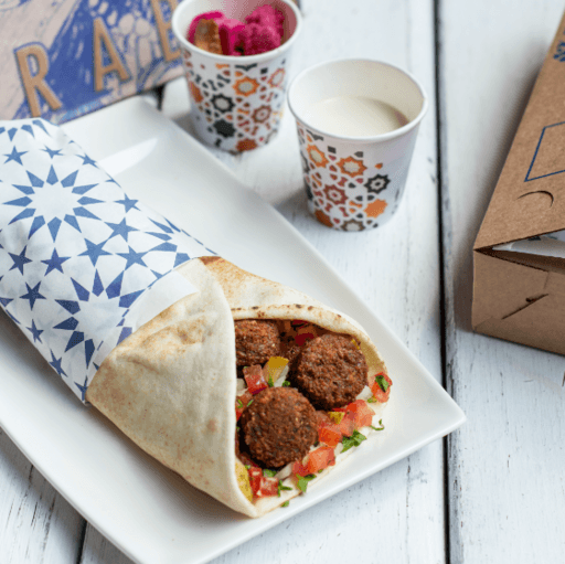 Vegano - Sanduíche de bolinhos fritos de fava e grão de bico com especiarias, cebola, tomate e salsinha picados, enrolados no pão árabe. Acompanha molho tarator e conservas.