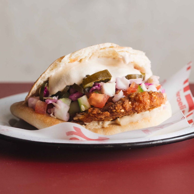 Filé de frango empanado com gergelim, salat israeli (tomates e pepinos em cubos), hummus, tehina, repolho e picles de pepino, servido na pita.
