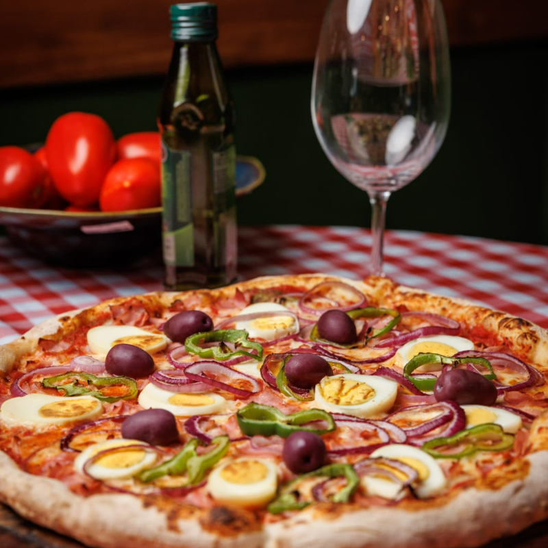 assa artesanal de fermentação lenta, molho de tomate artesanal, muçarela, presunto fatiado, rodelas de cebola roxa fresca, pimentão verde, ovos cozidos fatiados e azeitonas pretas inteiras. Finalizada com um fio de azeite extravirgem.