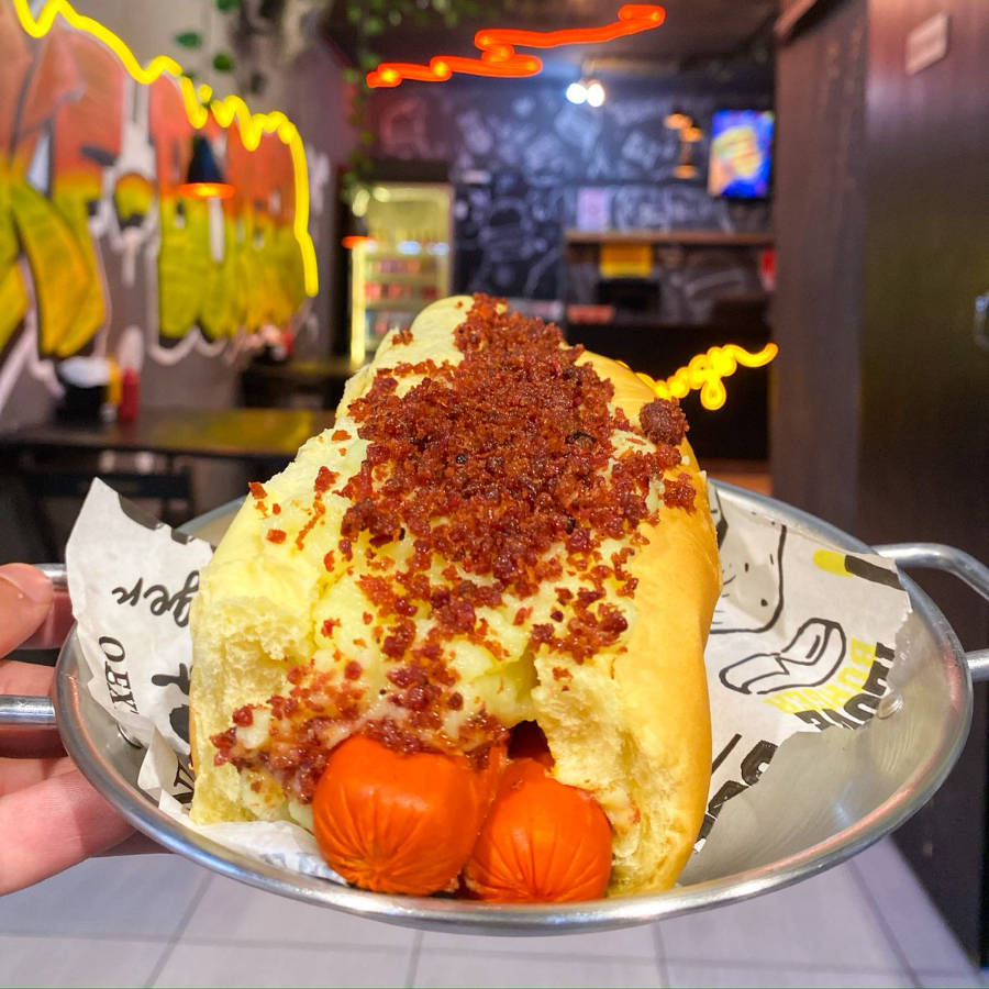 Pão brioche, 2 vinas (salsichas), maionese artesanal, ketchup, maionese artesanal, ketchup, mostarda, milho, tomate, bacon triturado, batata palha moída e purê de batata.