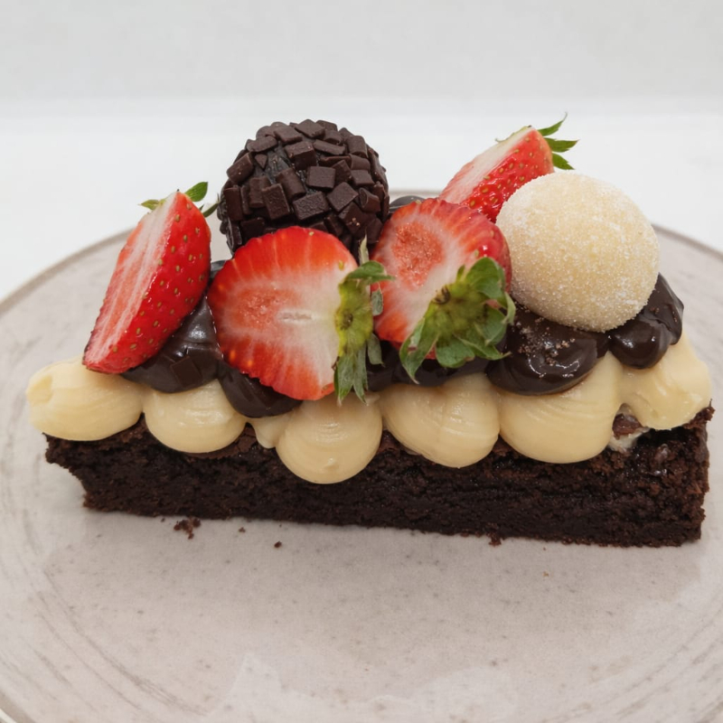 Fatia de brownie intenso, coberto com brigadeiro de leite ninho super cremoso, brigadeiro meio amargo, morangos frescos e bolinhas de brigadeirode ninho e chocolate. Uma combinação elegante, equilibrada e irresistível - doce na medida certa!