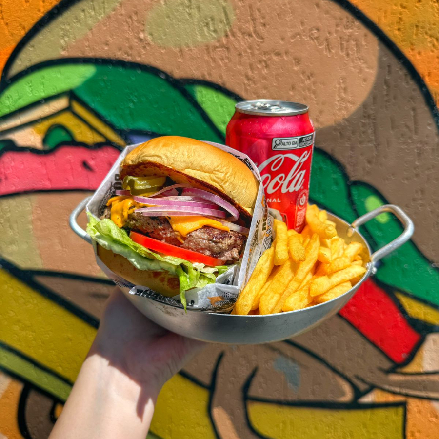 Pão brioche tostado na chapa com manteiga, hamburguer artesanal de aprox 160g, queijo cheddar fatiado, alface, tomate, picles, cebola roxa e maionese da casa. Porção de fritas aprox 150g (in natura) e uma lata de coca cola.