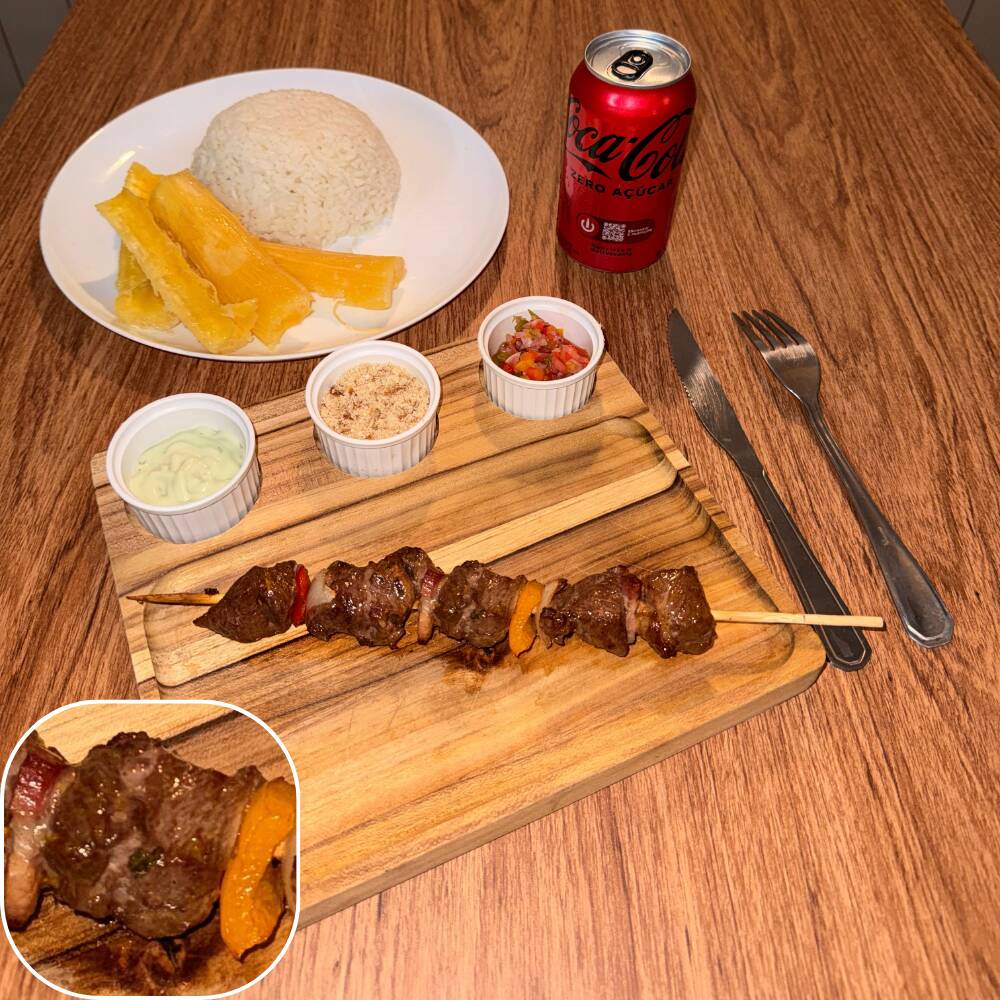 Espeto do miolo da alcatra bovina com cebola, bacon e pimentão acompanhado por arroz, mandioca, vinagrete, farofa crocante e molho verde.