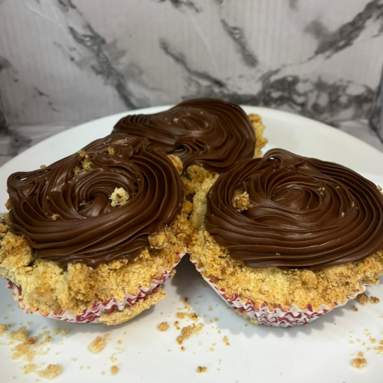 Muffin de chocolate no estilo cupcake, com massa bem fofinha e chocolatuda, recheio cremoso de chocolate e finalização generosa de chocolate ao leite por cima, coberto com uma farofinha amanteigada, docinha e levemente crocante. Uma explosão de textura e sabor em cada mordida! Dica da casa: aqueça de 15 a 20 segundos no micro-ondas até ficar quentinho e com o chocolate derretendo por dentro — o efeito é de bolo recém-saído do forno, simplesmente irresistível!