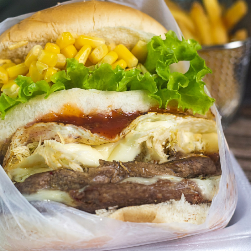 Desfrute do nosso X-Picanha, um dos sanduíches mais pedidos! É uma experiência inesquecível composta por um suculento hambúrguer de picanha de primeira qualidade, complementado pelo sabor intenso do presunto e queijo derretido. A adição do ovo e do bacon crocante traz uma explosão de sabor, enquanto o frango desfiado acrescenta uma textura única. O toque especial fica por conta do Catupiry cremoso e a batata palha crocante. Acrescentamos milho, alface fresca e tomate para um frescor equilibrado. Uma delícia que você vai adorar! Obs: Foto ilustrativa.