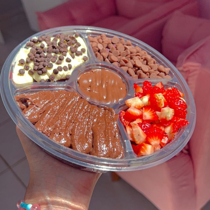 A roleta de açaí de 500ml com 5 adicionais é uma sobremesa servida em uma embalagem em formato de roda dividida em compartimentos, contendo açaí cremoso e cinco acompanhamentos à sua escolha.