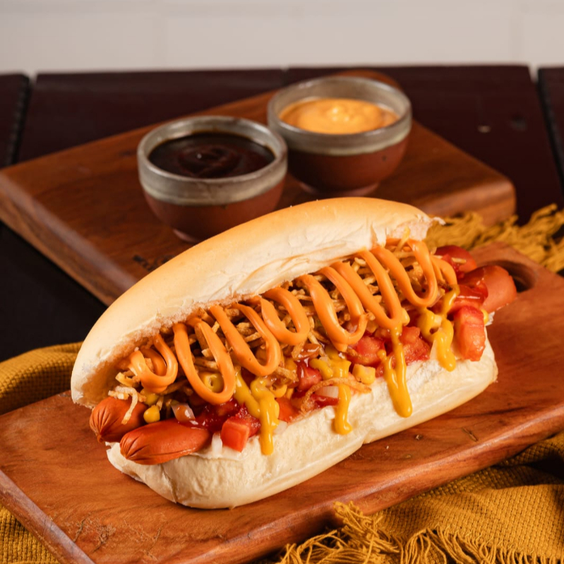 Pão prensado na chapa, 2 salsichas (PERDIGÃO), salada de tomate e cebola frescas, milho, maionese, batata palha, ketchup, mostarda e para finalizar cheddar cremoso.