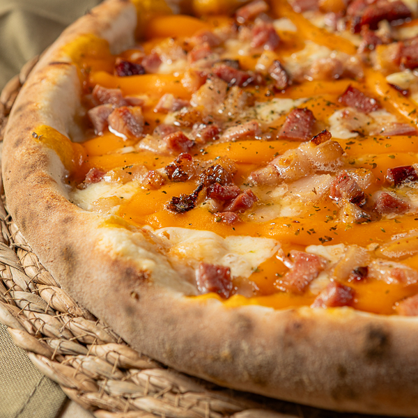 Na pizza de cheddar e bacon vai: molho de tomate, queijo ralado mussarela, tomate, cheddar, cebola, bacon e orégano