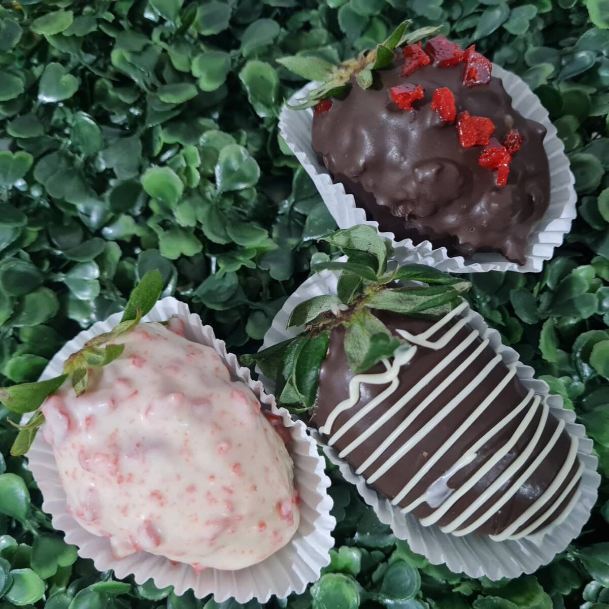 Três maravilhosos doces com morango fresco envolvido por brigadeiro / brigadeiro branco e cobertos por chocolate / chocolate branco e e pedaços crocantes de caramelo vermelho.