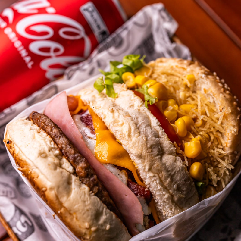 X TUDO( Pão artesanal, carne de hambúrguer bovino, queijo prato derretido, presunto, ovo frito, bacon e m cubos, batata palha fina artesanal, milho verde, alface crespa e tomate fresco) acompanha coca cola lata