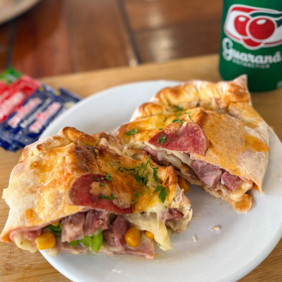 Calzone À Moda feito com massa leve e crocante, assado na medida certa e recheado com uma combinação irresistível de muçarela, presunto, bacon, calabresa, pimentão, cebola, tomate, milho e orégano. Bem recheado, suculento e perfeito pra matar a fome de verdade!