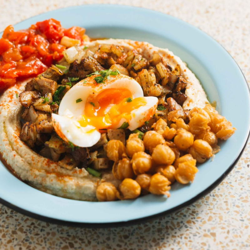 Hummus, coxa e sobrecoxa temperadas com cebola na chapa, grão-de- bico crocante, ovo mole, matbucha, azeite e páprica.