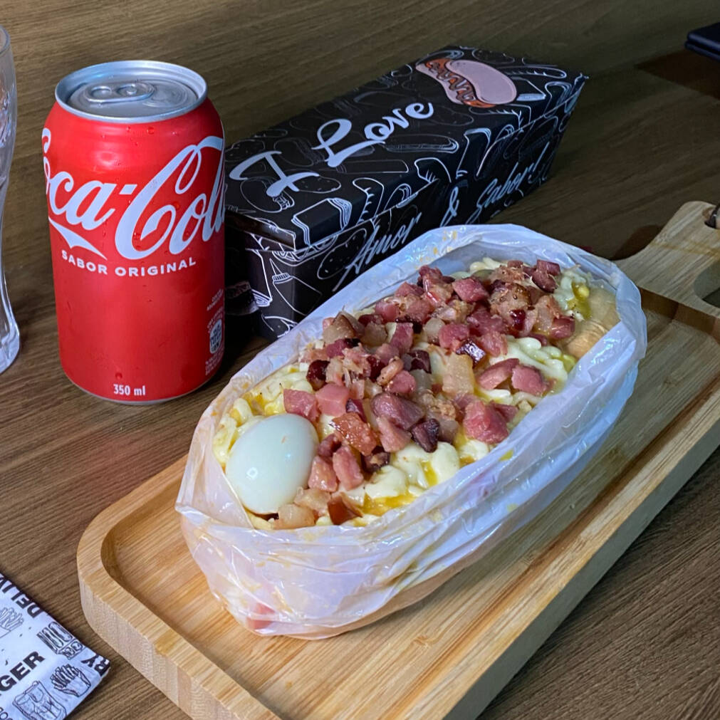 Acompanha: Pão, Salsicha ou Linguiça, Molho, Milho, Ervilha, Molho da Casa, Batata Palha, Cheddar ou Catupiry, Queijo Mussarela Gratinada, Bacon e Ovo de Codorna. *Coca-cola lata da imagem não inclusa. Pedir separadamente na categoria "bebidas".