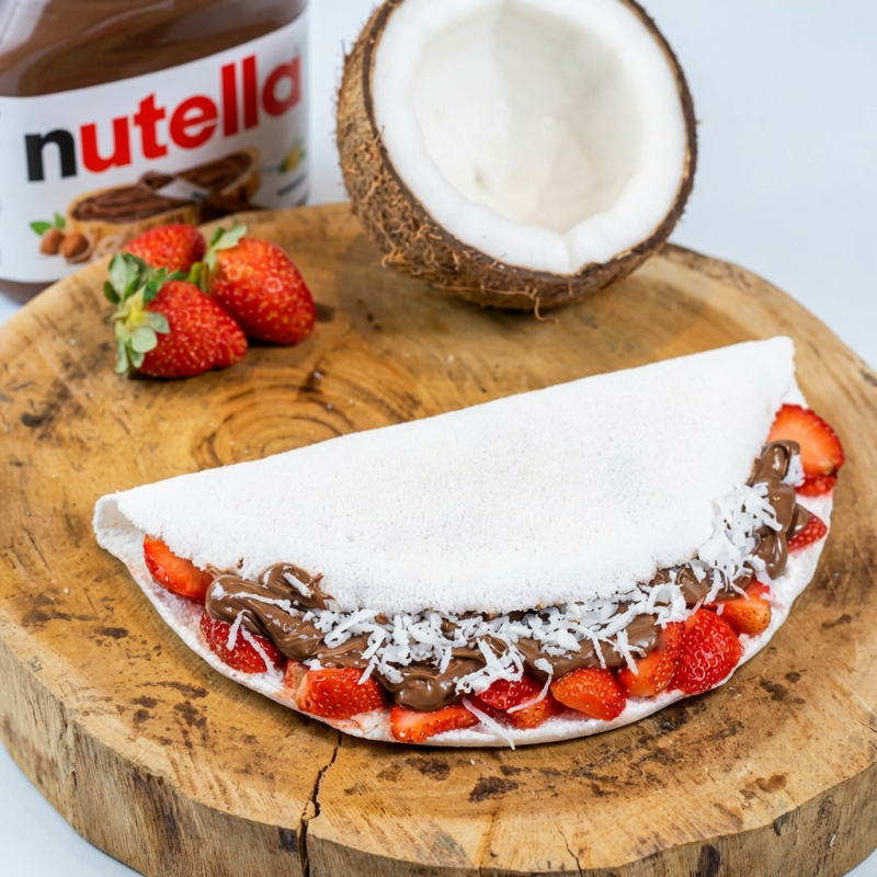 A 'Explosão de Sabor' é uma estrela da nossa categoria 'Tapiocas Doces'. Esta harmonia de sabores combina morangos frescos e suculentos com a indulgência da Nutella. Tudo é delicadamente envolvido por uma tapioca leve e macia. O coco fresco ralado finaliza com uma textura interessante e doçura tropical. Prepare-se para uma experiência culinária memorável, onde cada mordida é uma verdadeira explosão de sabor!