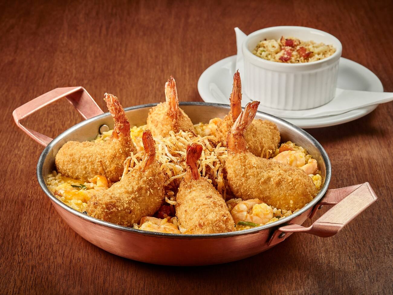 Camarões empanados e recheados com Catupiry, sobre um cremoso arroz de moqueca com camarões e coentro, coberto com batata palha. Acompanha farofa de panko com bacon e alho poró.