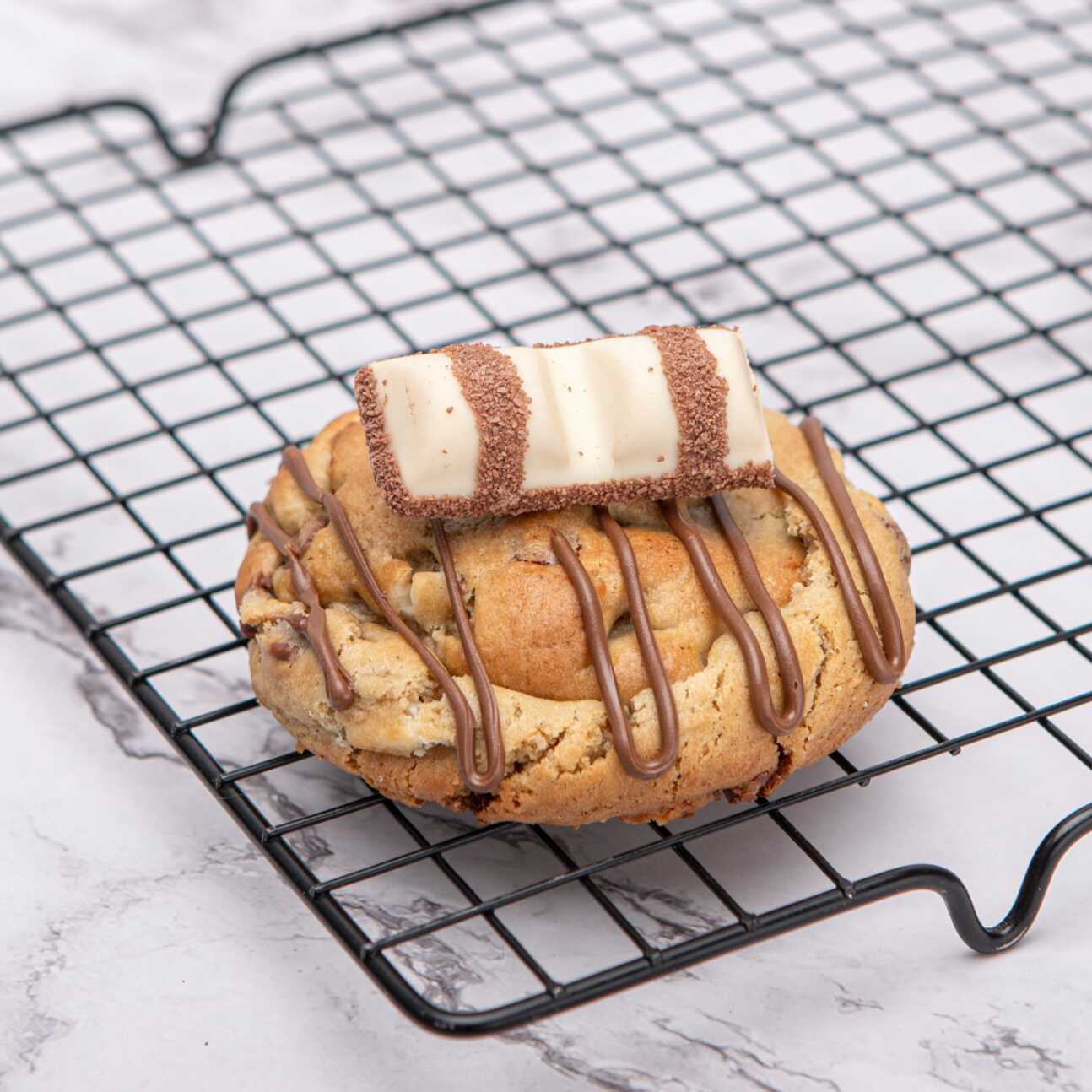 Um cookie irresistível, com uma massa amanteigada com gotas de chocolate ao leite e chocolate branco, recheado com um recheio cremoso inspirado no Kinder Bueno, finalizado com creme de avelã e Kinder Bueno.