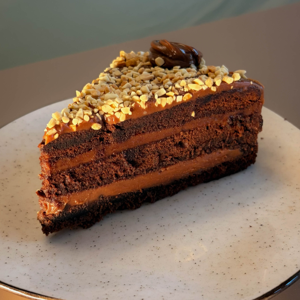 Fatia generosa de bolo inspirado no sabor de Ferrero Rocher, recheio de creme de avelã e Nutella. Com um bolo de chocolate molhado feito com cacau puro. No topo tem cobertura de creme de avelã, e amendoim triturado, finalizado com uma decoração de Nutella com folhas de ouro. Aprox: 250g