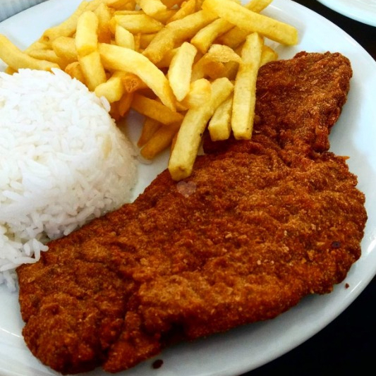Á milanesa de bife contra filé empanado na farinha de rosca, arroz, feijão e batata frita.