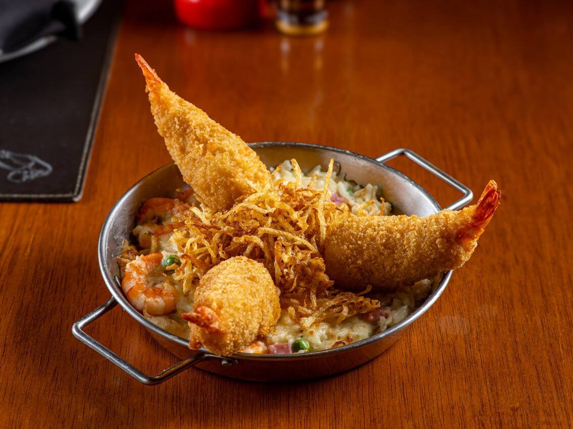 Camarões (30g/cada) recheados com Catupiry e empanados. Servidos sobre cremoso arroz com camarões (18g/cada), ervilhas e presunto, envolvido com molho branco. Gratinado com queijos muçarela e parmesão. Acompanha batata palha. Tamanho do camarão nesse