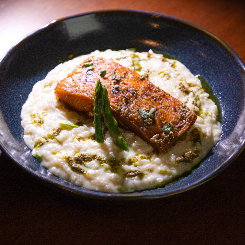 Posta de salmão, acompanha base de arroz arbóreo com vinho branco seco e parmesão. Finalizado com aspargos e molho pesto.