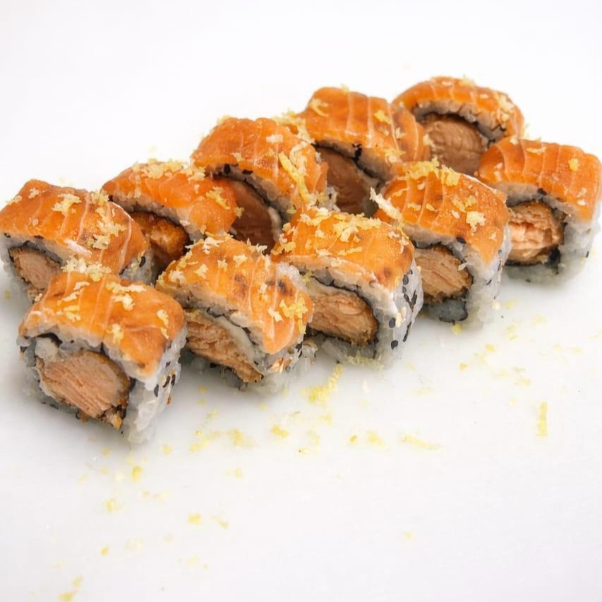 Uramaki com salmão empanado no recheio, coberto com fina fatia de salmão cru e finalizado com raspas de limão siciliano, trazendo um toque cítrico e refrescante que realça o sabor do peixe.