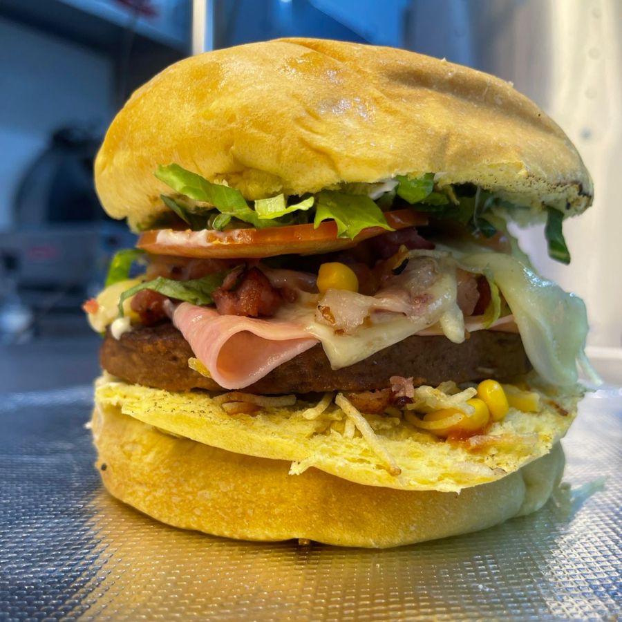 Pão prensado, hambúrguer, mussarela, presunto, bacon, ketchup, maionese, alface, tomate, batata palha e milho.