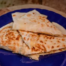 A 'Quesadilla de Carne - La Catrina' recebe esse nome em homenagem ao Dia dos Mortos mexicano, 'Los Dias de los Muertos'. Este prato consiste em uma deliciosa tortilha de trigo crocante e dourada, habilmente cortada em quatro generosas fatias. Cada seção é primorosamente recheada com um mix de queijos derretidos, carregados de sabor e cremosidade. Complementando sua riqueza, a carne desfiada, macia e suculenta, se une aos tomates frescos e suculentos, proporcionando um equilíbrio perfeito de sabores. Esta quesadilla é uma autêntica celebração da culinária mexicana em cada mordida.