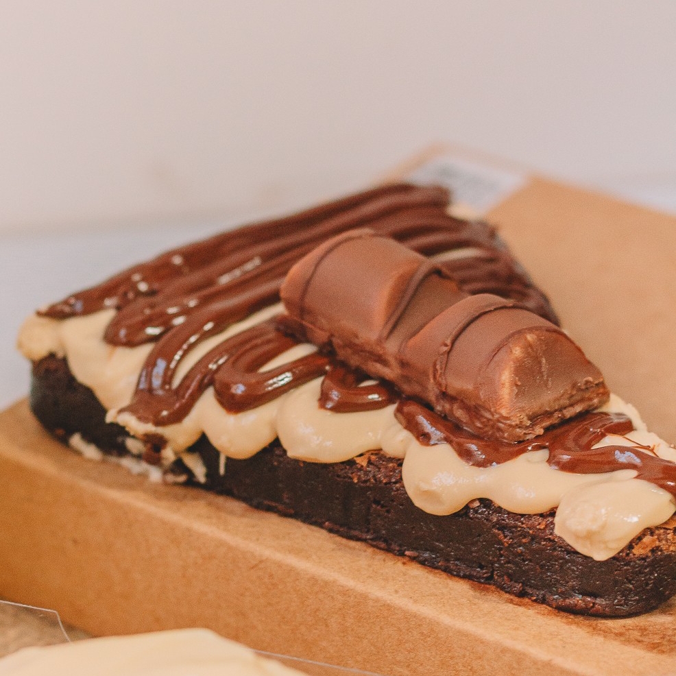 Uma base irresistível de brownie denso e úmido, preparado com chocolate nobre que derrete na boca. No centro, um creme cremoso inspirado no Kinder Bueno traz doçura equilibrada e textura aveludada. Para finalizar, uma camada generosa de Nutella se espalha brilhante por cima, coroada com um Kinder Bueno que adiciona crocância e aquele sabor inconfundível.