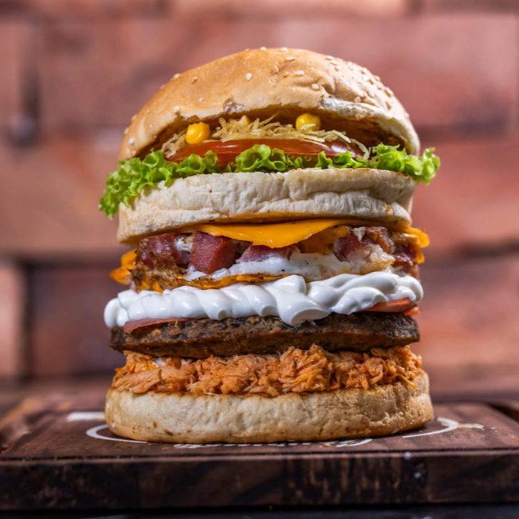 Hambúrguer (Pão artesanal, carne de hambúrguer bovino, queijo prato derretido, presunto, ovo frito, bacon em cubos, frango desfiado, catupiry cremoso, batata palha fina, milho verde, alface crespa e tomate fresco)