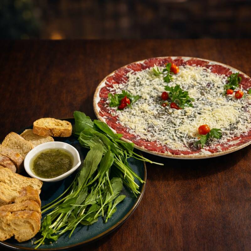 Sofisticado na medida certa e cheio de sabor! Finas fatias de carpaccio, macias e delicadas, servidas com salada de rúcula fresca que traz aquele toque levemente picante e aromático. Finalizado com lascas de parmesão que acrescentam intensidade e textura, além de molhos especiais que envolvem cada ingrediente com equilíbrio perfeito. Acompanha torradas crocantes, ideais para montar cada pedaço e aproveitar essa combinação clássica e irresistível. Uma entrada elegante, leve e perfeita para começar a refeição com estilo.