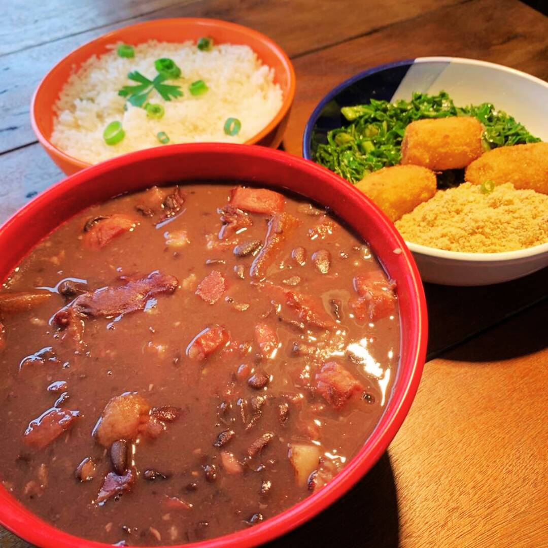 Feijoada completa e bem servida, ideal para dividir. Preparada com carnes selecionadas: costelinha suína, paio, calabresa, bacon, joelho suíno, pé salgado, máscara suína salgada e charque, com cortes bovinos e suínos cozidos lentamente para garantir muito sabor. O kit acompanha uma marmita média apenas com feijoada, uma marmita pequena de arroz branco e uma marmita pequena com farofa, couve refogada e banana frita. Uma refeição tradicional, completa e muito saborosa. Serve até 2 pessoas.