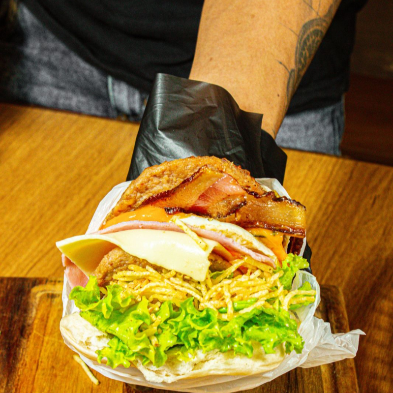 Pão, 3 carnes, 1 ovo, bacon, cheddar cremoso, queijo, presunto, alface e batata palha.