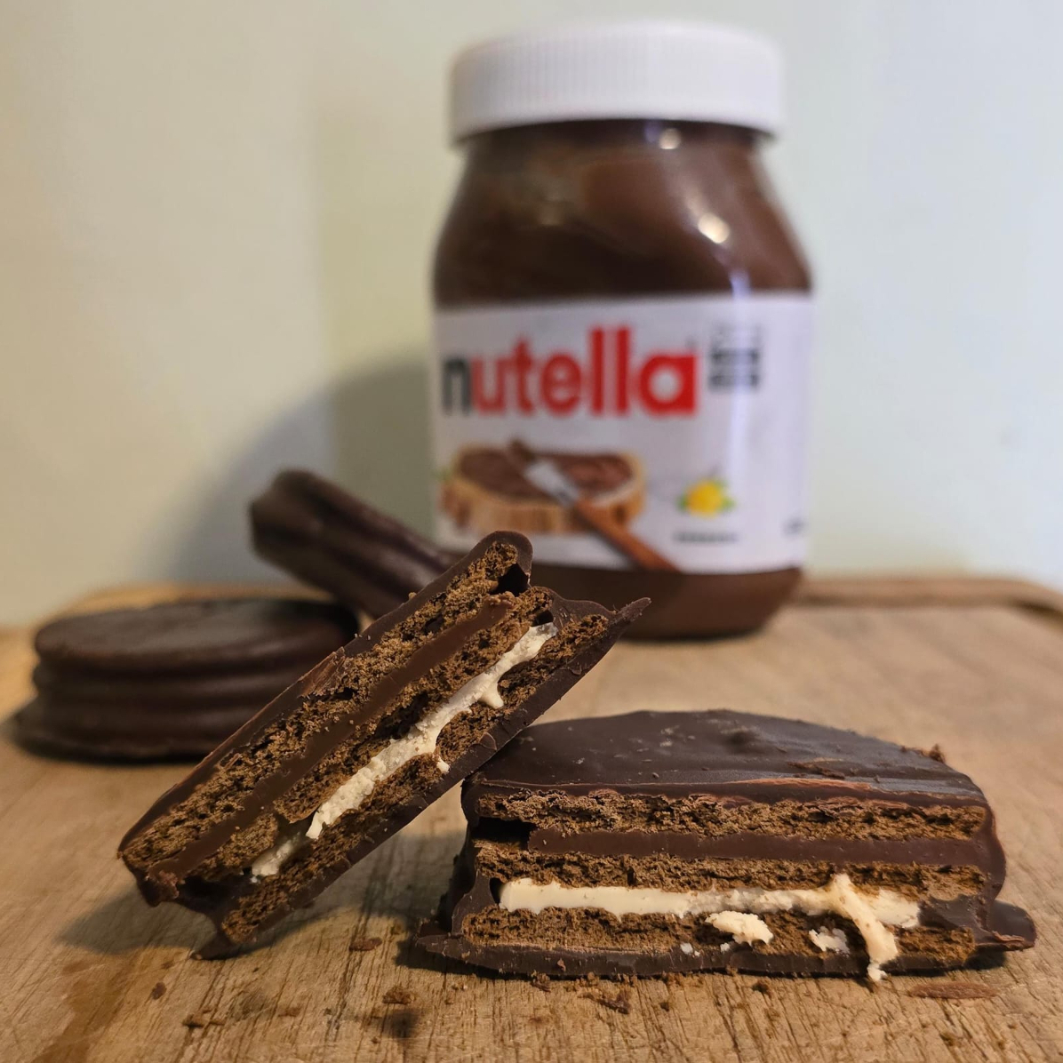 Alfajor rústico e artesanal com biscoito especial crocante, uma cama de recheio de Nutella pura, uma camada de creme tipo Kinder Bueno e uma casquinha delicada de chocolate meio amargo.