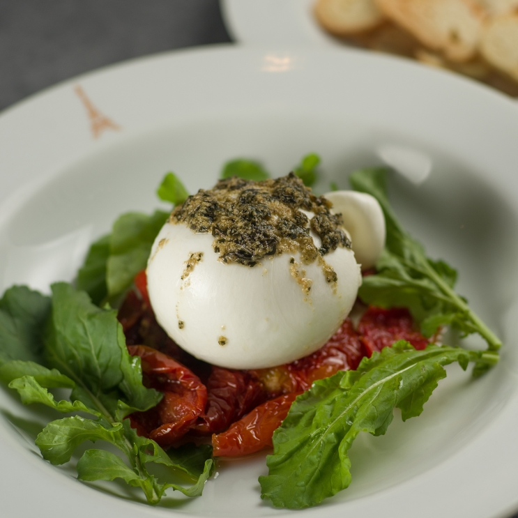 Nossa refinada e cremosa burrata de búfala artesanal, servida sobre uma generosa cama de tomate confit, regada com molho pesto e acompanhada de torradas crocantes.